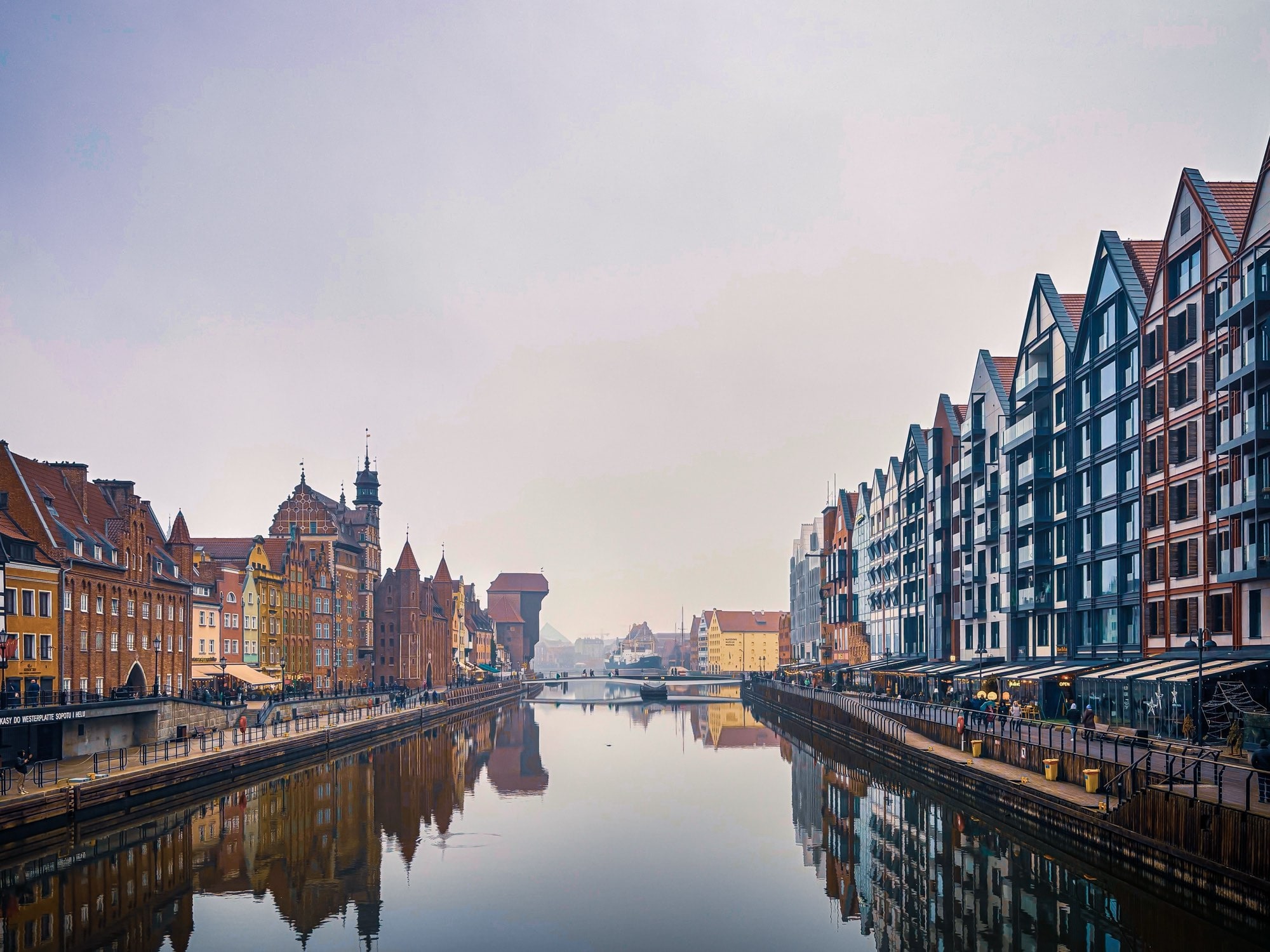 The Motława seen from a Gdańsk bridge on a still, hazy morning, granary gables and Old Town roofs doubled in the flat water.